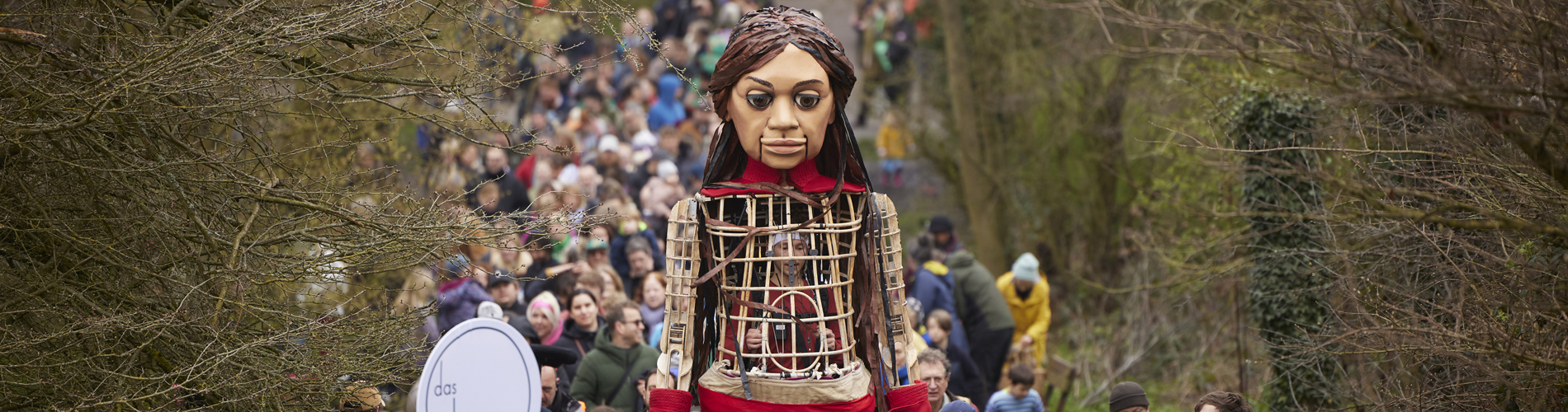A crowd of people walk with Little Amal, a large 12 foot puppet of a brown haired child.