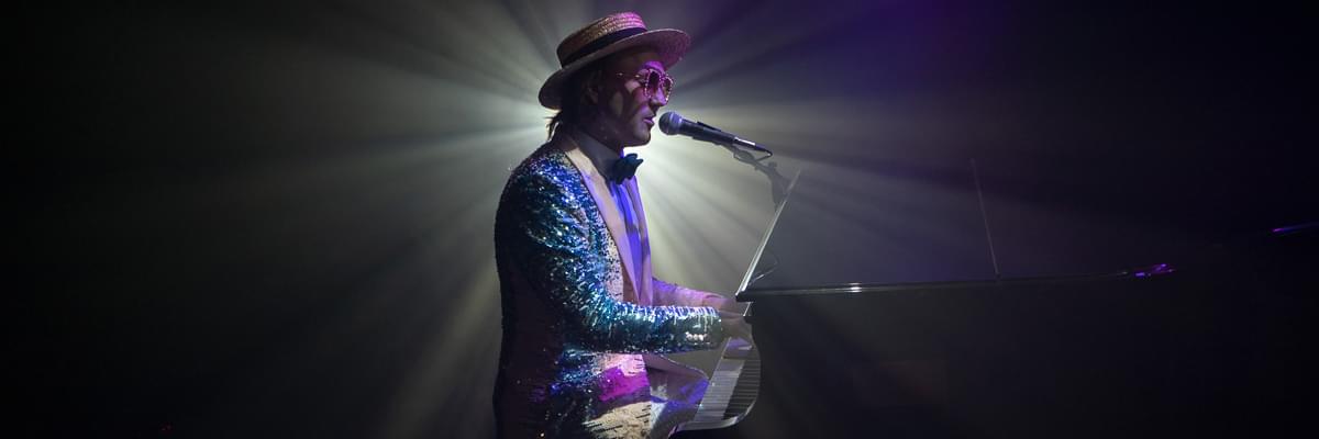 A performer dressed as Elton John sets at a black grand piano, playing and singing into a microphone. He wears a sparkly jacket and glasses and a straw boater hat. A strong spotlight shines directly behind him.