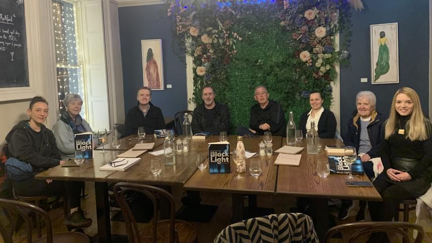 Eight people sit around 4 long tables pushed together in a high ceilinged room. Three books titled 'Blacklight' are propped up on the tables, as well as bottles and glasses of water.