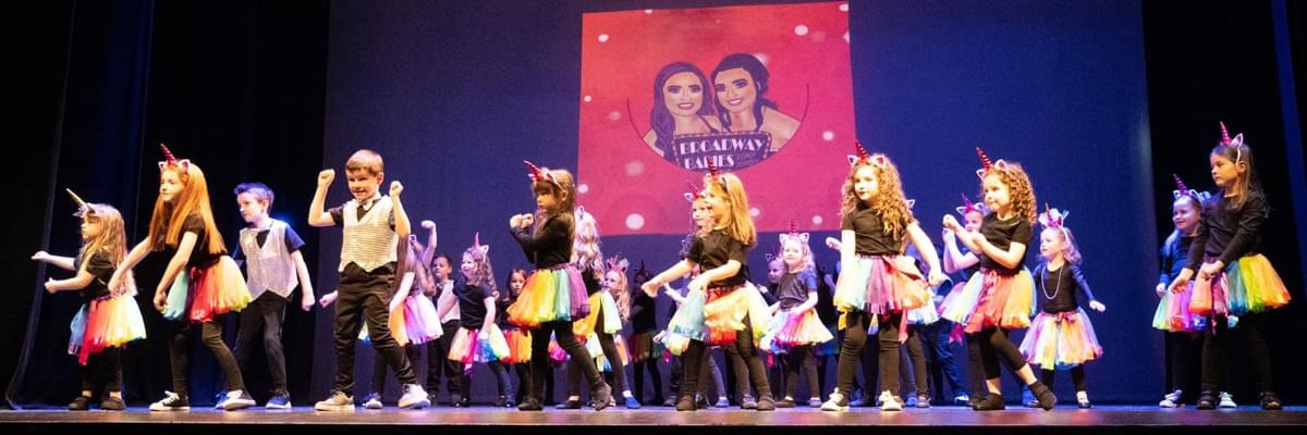 Mostly young girls in black tshirts and rainbow tutus on a high stage.