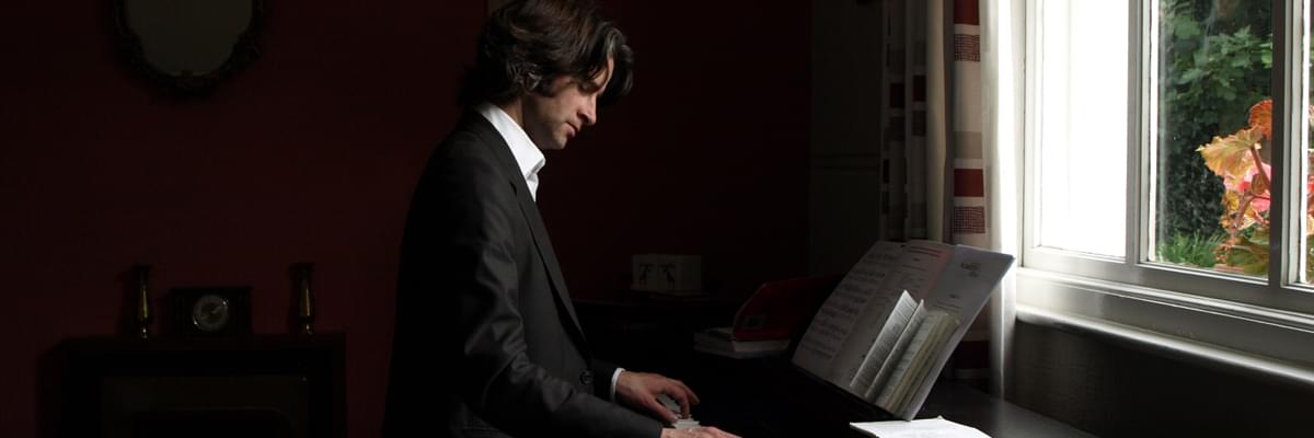Man with longer hair in a black suit with white shirt, in profile facing a window. He's looking down with his hands on piano keys.