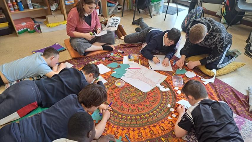 A woman sits on the floor with students lying in a circle drawing picitures