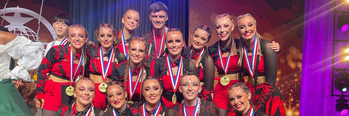 Group of smiling young people face the camera, all wearing matching red and black dance outfits, each with a medal around their necks.