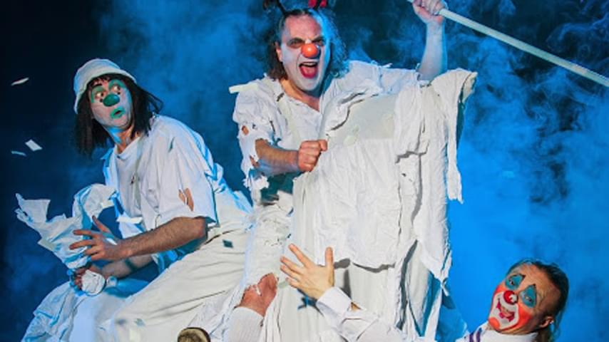 Three clowns face the camera dressed in various outfits made of plain, white paper and tshirts, with a smoky, blue background behind them.