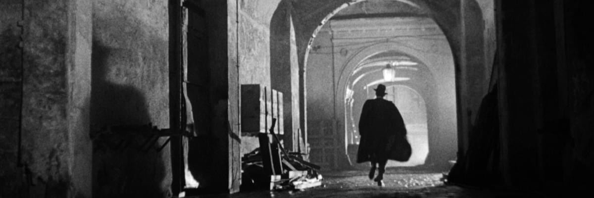 Black and white photo of a dark, curved bricked tunnel, lit from the end of the tunnel. A man in a wide brimmed hat and dark cloak runs away from the camera