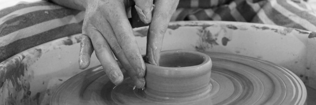Black and white photo of a person in a striped apron sitting over a pottery wheel, crafting a small pot with wet clay.