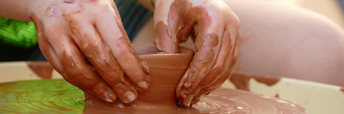 Young person sits over a pottery wheel, crafting a small pot with wet clay.