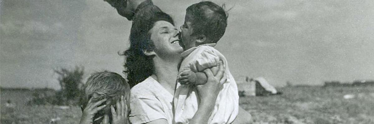 Black and white, vintage looking photo of a beach scene, of a mother holding her baby, smiling, with two young boys next to her.