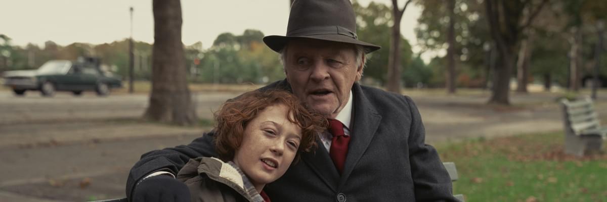 A young boy with wavy red hair, sits on a park bench, head on the shoulder of a older man, played by actor Anthony Hopkins.