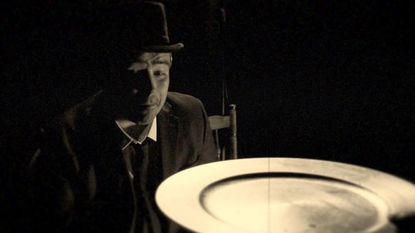 Sepia toned photo - Member of Encore Productions sits in a wooden chair, dressed in a white shirt, dark suit jacket, tie and hat. They look at a shiny metallic plate.