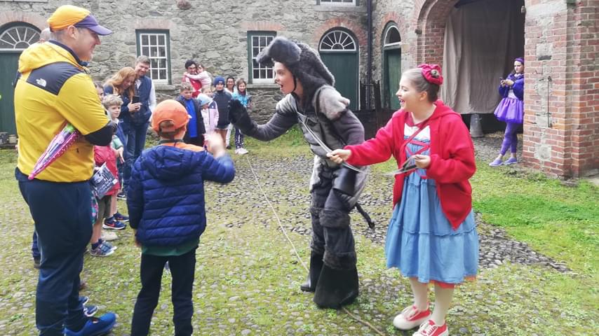In an old style brick courtyard, a family audience to the left looks at three actors to the right. The actors are dressed, one in head to toe bright purple, one in a denim dress and red hoodie, and one in a full donkey costume. The actor in the donkey costume is giving one of the child members of a the audience a high five.