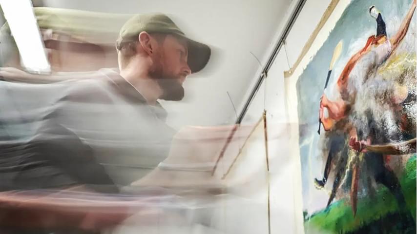 Action shot of a young man with a beard and baseball cap, painting onto a canvas on a wall
