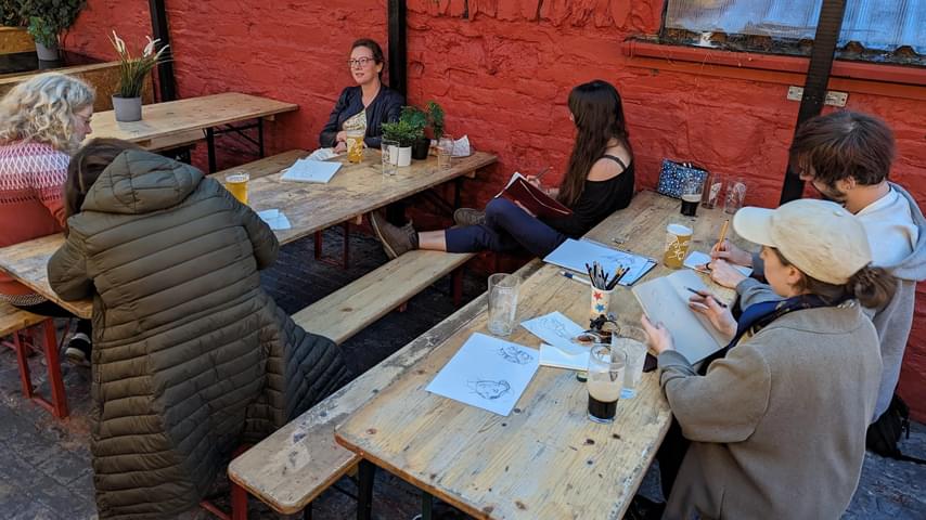 Several people are sat around 2 picnic style tables in a beer garden area in the pub Mó Chara, Dundalk, Co. Louth. They are sketching in notebooks, and there are drinks on the tables.