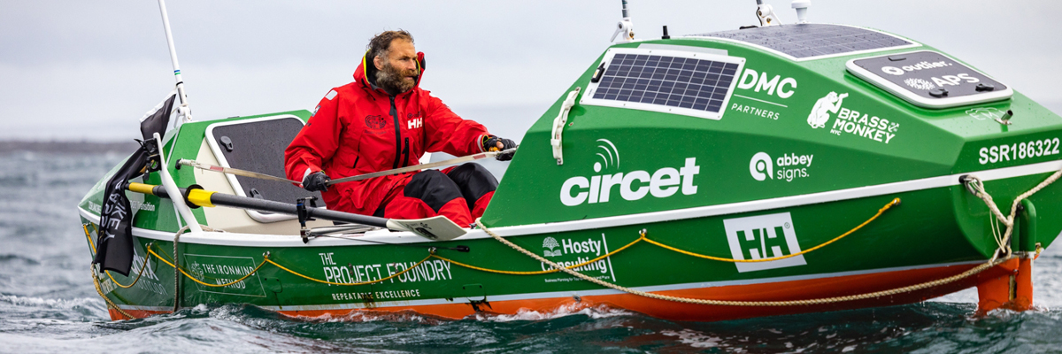 A bearded man sits in a hi-tech looking large green row bow in the ocean. He wears red weather proof overalls.