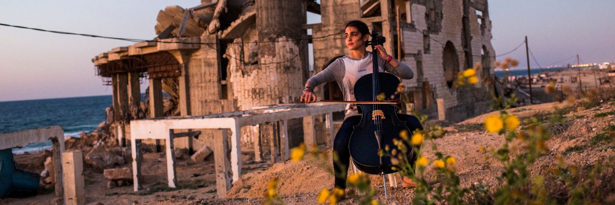 A girl plays a cello in the rubble of a bombed building in Gaza against a hazy sky.