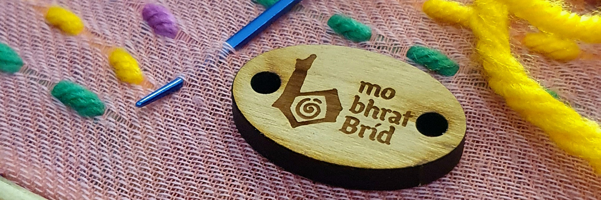 Embroidery loop with pink fabric and stitched with turquoise, yellow and purple wool. A wooden engraved oval lays on top witht he words Mo bhrat Bríd