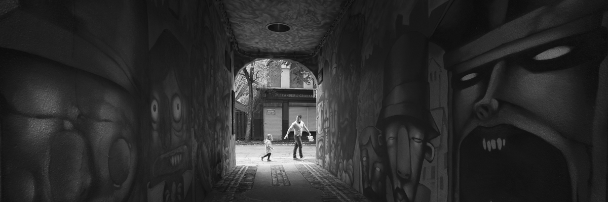 Photo of a dark, graffitied tunnel, at the end of the tunnel are a father and child walking together.