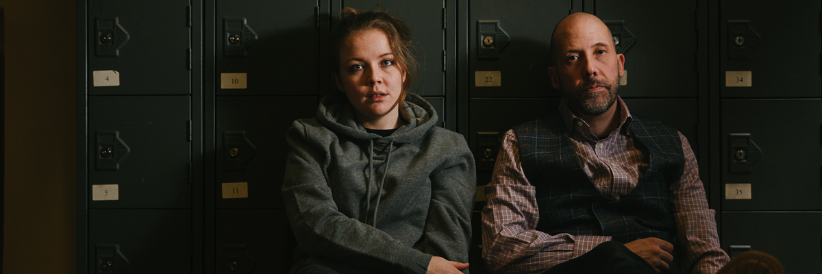 A young woman, wearing a grey hoodie and older man, wearing a shirt and waistcoat, sit on the floor against lockers.
