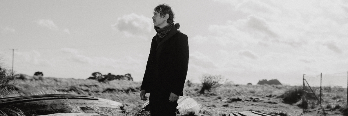 Black and white photo of a man with a long black coat and scarf standing in a grassy outdoor scene