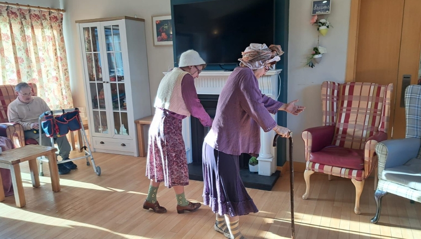 Two ladies dressed in purple and wearing white hats walk with walking sticks