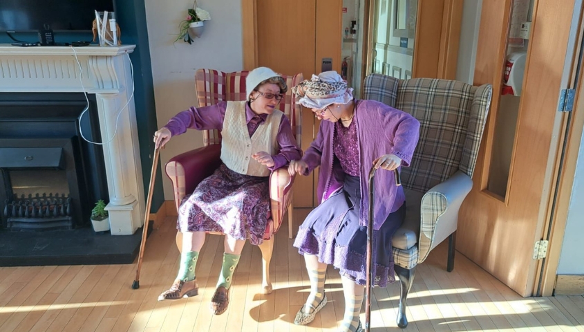 Two Ladies dressed in purple and white sit in armchairs holding walking sticks