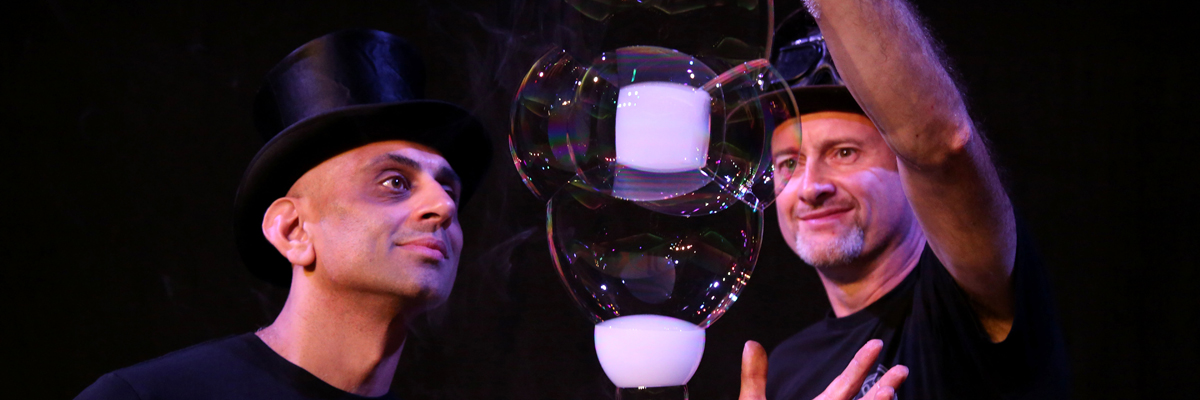 Two men in top hats and black tshirt, one is holding up a chain of large bubbles