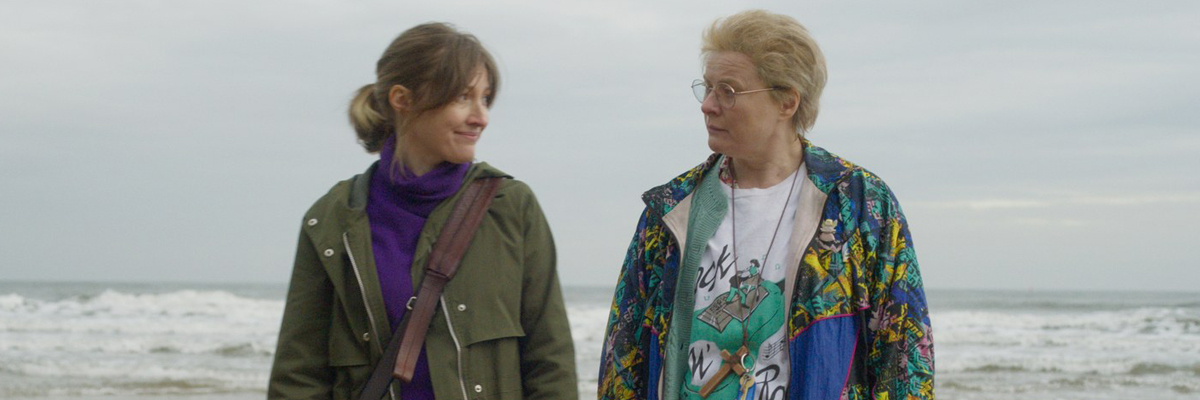Two women, one younger, one older, stand on a beach facing the camera but heads turned to face each other.