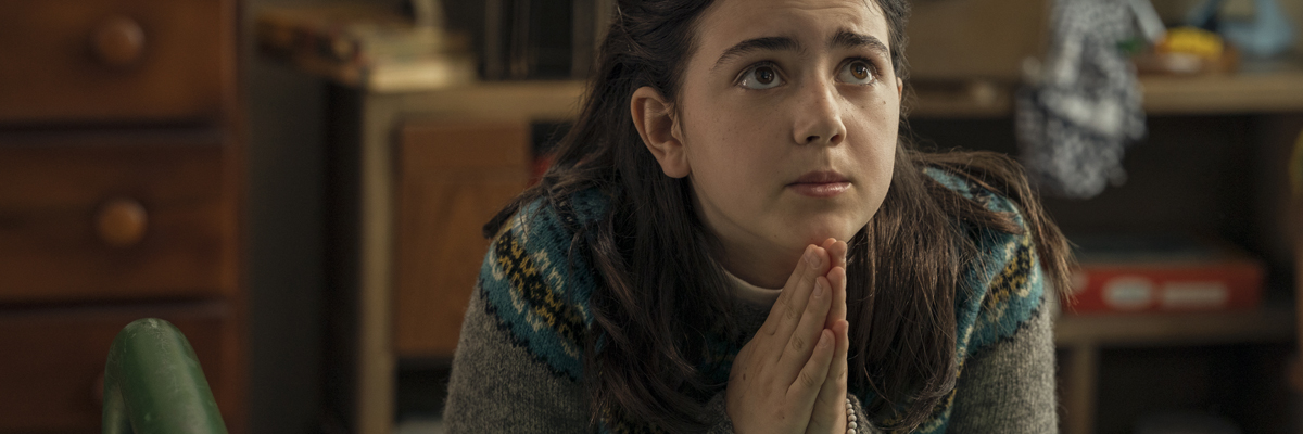 A young girl with long dark hair and 70s style sweater leans on her bed with her hands together in prayer.