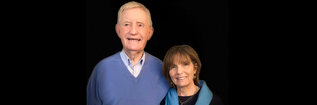 A older man and woman stand facing the camera, smiling. The man wears a blue sweater, the lady a dark blue sweater and bright blue scarf