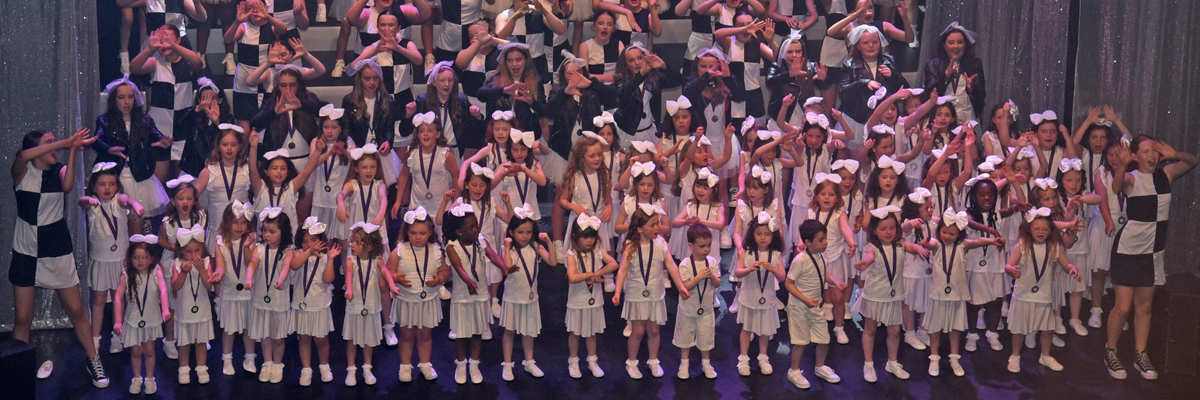 A large group of children aged between approx. 5 to 15 years old, dressed in black and white outfits are performing a dance on a large stage