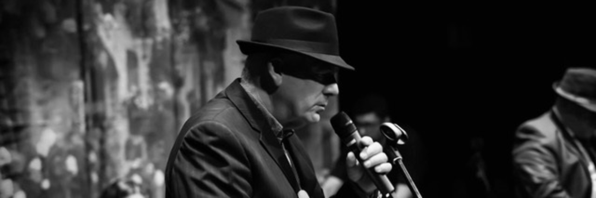 A dark photo of a man on stage, leaning into a microphone. He wears a dark suit and hat.