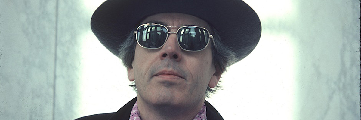 A man's head and shoulders are visible, he wears sunglasses, a wide brimmed hat, pink shirt and dark jacket