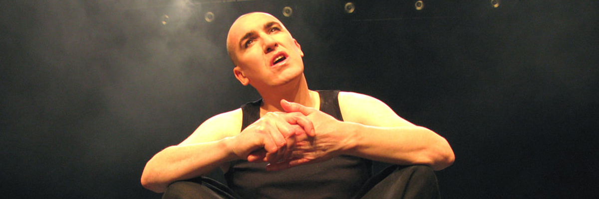 A bald man sits crossed legged on a stage. He wear a black tank top and sweatpants