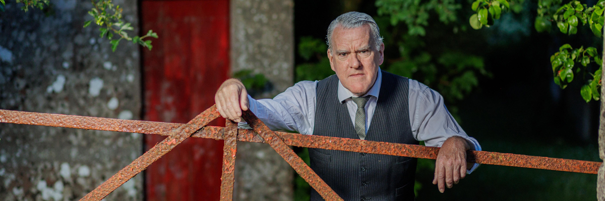 A man with grey hair and eyebrows lends on a metal gate. He wears a light shirt, grey waistcoat and green tie.