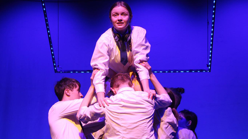 A small group of students in white shirts and ties hold up a female student above them. The background is a vivid purple.