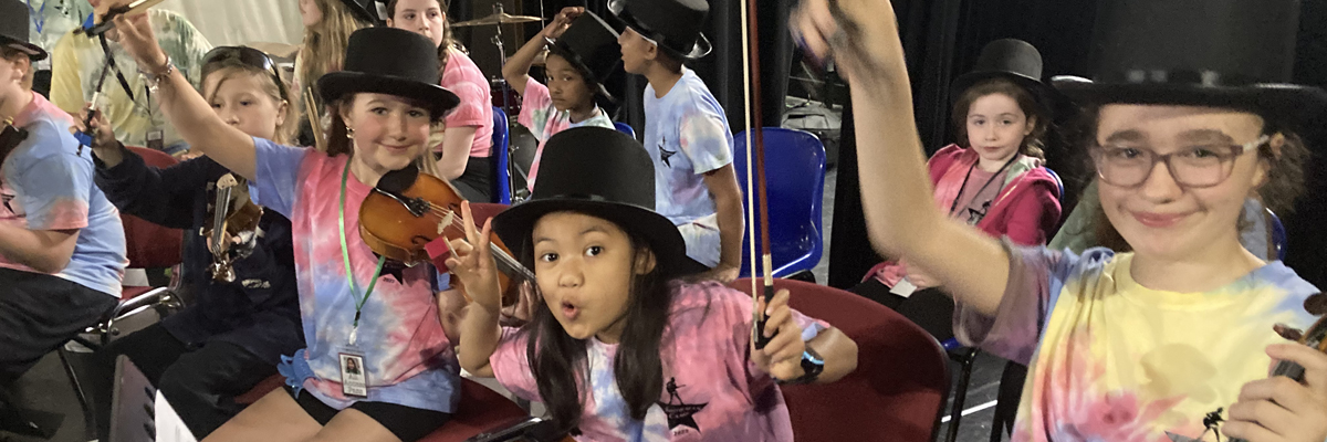 Children smile and wave at the camera, They are sitting, wearing tie dye shirts and top hats and holding instruments.