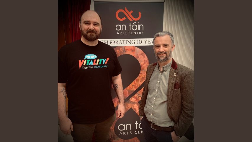 Two men with beards dressed casually stand in front of an An Táin Arts Centre Pop Up.