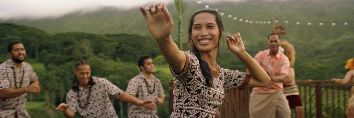 A Samoa lady smiles and dances. Other Samoa people and a beautiful green mountain are behind her.