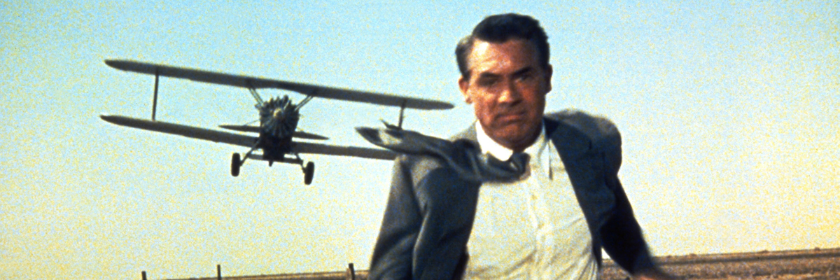 An old colour photo of a man in a gray suit and white shirt running while a propeller plane can be seen flying low over his shoulder.