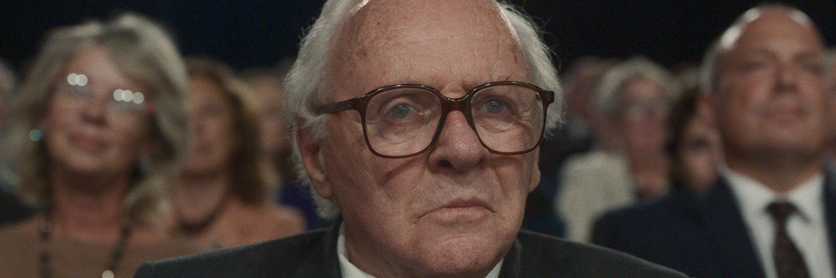 An older man wearing glasses sits in a crowded theatre looking forward intensly.