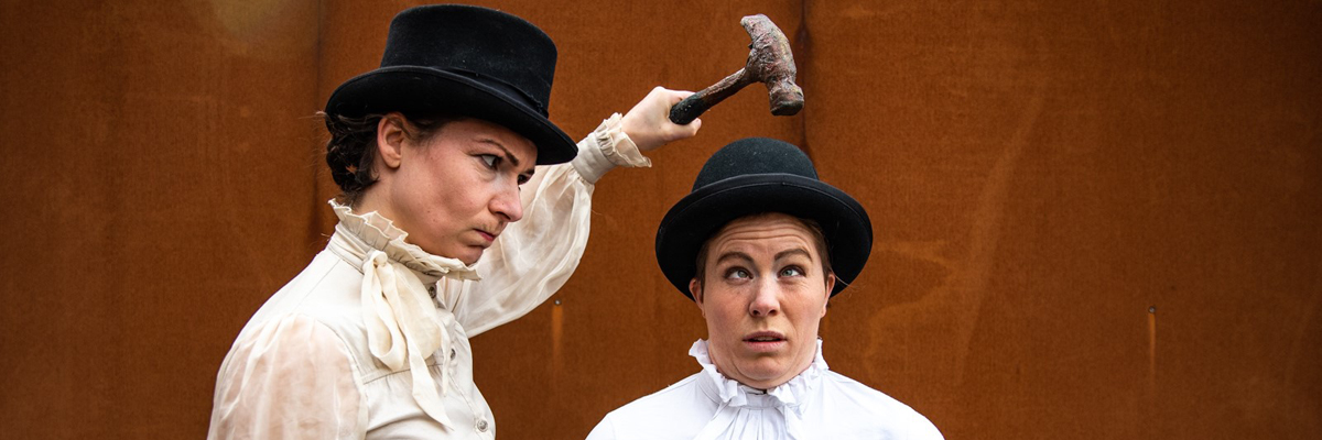 Two women dressed in old fashioned, high collared white shirts and bowler hats. One holds a hammer above the others head.